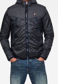 Man som bär en svart zip-up huva jacka med texturerade axlar och blå jeans, med händerna i fickorna, stående mot en neutral bakgrund.