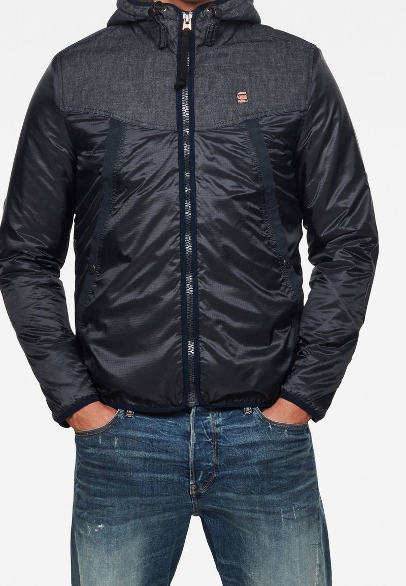 Man som bär en svart zip-up huva jacka med texturerade axlar och blå jeans, med händerna i fickorna, stående mot en neutral bakgrund.