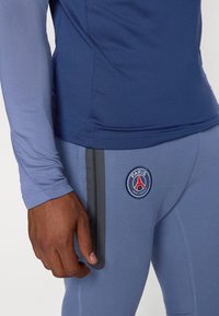 Marineblå sportsbluse med lange ærmer, parret med lyseblå tætsiddende bukser, med Paris Saint-Germain logo og grå accentstribe på siden.
