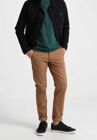 Mann steht mit Händen in den Taschen, trägt eine schwarze Jacke, ein grünes Hemd, beigefarbene Hosen und schwarze Sneakers vor weißem Hintergrund.