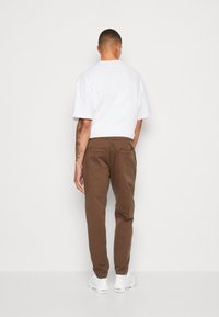 Braune Baumwollhosen mit lockerer Passform, ausgestattet mit zwei Gesäßtaschen. Kombiniert mit einem schlichten weißen T-Shirt und weißen Turnschuhen.