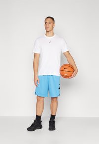 Bela majica s kratkimi rokavi z črnim logotipom, modri košarkarski šortsi, črne superge, drži oranžno košarko. Preprost dizajn, atletski kroj.