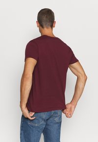 Man met kort bruin haar in een effen bordeauxrode t-shirt met korte mouwen en blauwe jeans, staand met zijn rug naar de camera tegen een witte achtergrond.
