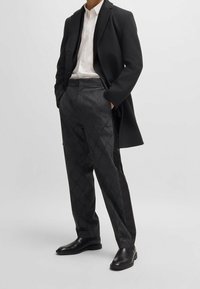 Cappotto lungo nero di lana indossato su una camicia bianca, abbinato a pantaloni neri a fantasia e stivali alla caviglia neri e eleganti. Motivi geometrici dettagliati sui pantaloni.