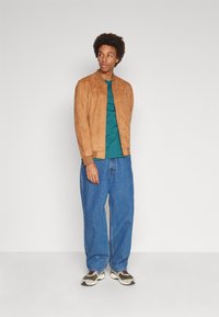 Jacket bomber in suede tan, t-shirt verde, jeans blu in denim larghi e sneakers multicolori. Design semplice con una vestibilità rilassata.
