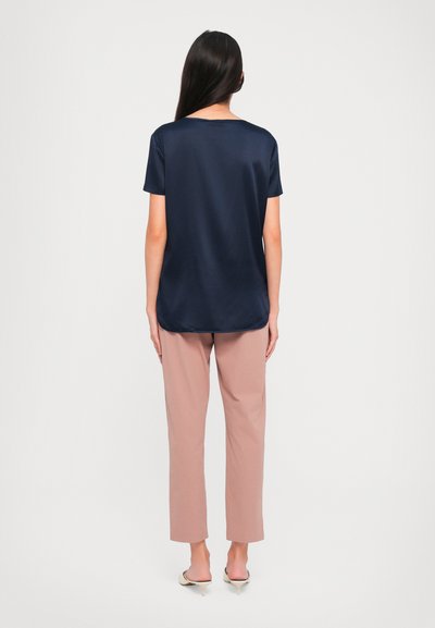 Woman with long dark hair wearing a navy satin short-sleeve blouse, light brown straight pants, and beige heeled mules, standing facing away.