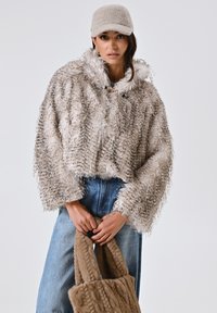 Giacca cropped in pelliccia sintetica nei toni beige e marrone, con cappuccio e maniche larghe. Abbinata a pantaloni in denim blu chiaro e una borsa a maglia marrone.