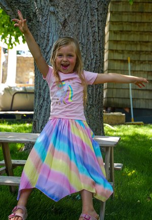 Mädchen trägt ein pinkes T-Shirt mit einem Einhorn-Motiv und einen mehrfarbigen, gestreiften Rock in Pastellfarben. Sie steht auf einer Holzbank im Freien.