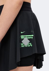 Άτομο που φοράει μαύρη φούστα Nike Running με νέον πράσινο κείμενο, ασημί ανακλαστικό τετράγωνο και ορατές συντεταγμένες στο αριστερό ισχίο.