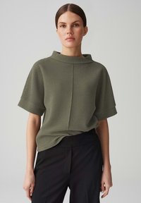 Top verde oliva a maniche corte con collo alto, tessuto testurizzato e vestibilità rilassata, abbinato a pantaloni neri.