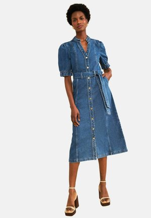 Naine seisab ja kannab keskmise pikkusega sinist teksakleiti õhuliste varrukate, eessete nööpide ja sobiva vööga, paaritatud beežide platvormkontsadega.