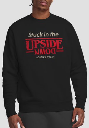 Black sweatshirt worn by a man, featuring text "Stuck in the UPSIDE DOWN *Since 1983*" with "UPSIDE DOWN" in large red letters upside down.