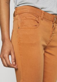 Le jean en denim orange présente une coupe ajustée, une fermeture éclair apparente et des accents métalliques. Le tissu a une texture douce avec des détails de couture subtils.