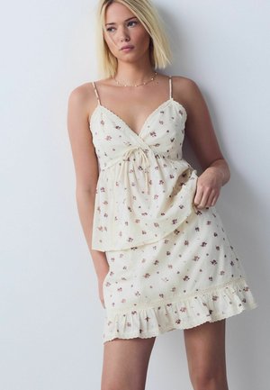 Młoda kobieta o blond włosach ubrana w kremową, kwiecistą sukienkę bez rękawów z koronkową lamówką, stoi na tle jednolitej jasnej ściany, trzymając brzeg sukienki.