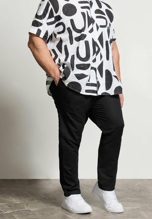 Mann steht mit den Händen in den Taschen, trägt ein weißes Hemd mit schwarzen abstrakten Formen, schwarze Hose und weiße Sneakers auf einem gefliesten Boden.