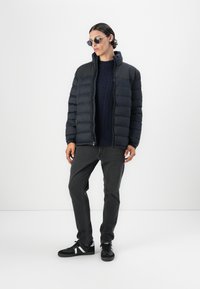 Puffer jacket in black with quilted design, high collar, paired with a navy sweater, gray jeans, and black sneakers featuring white accents.