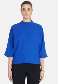 Blå, löst sittande topp med hög halsringning, trekvartsärmar, tillverkad av ett slätt tyg. Bärs med svarta byxor för kontrast.