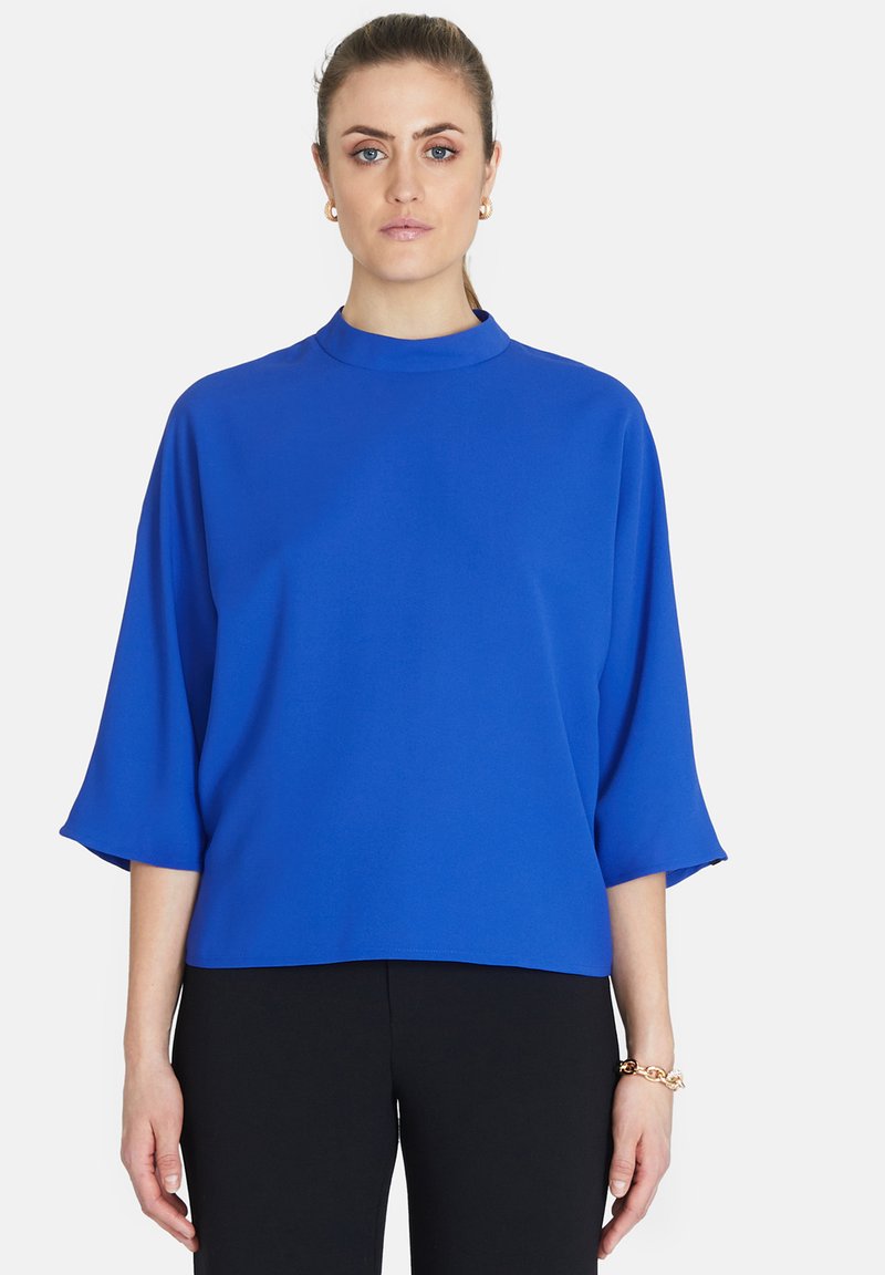 Blå, löst sittande topp med hög halsringning, trekvartsärmar, tillverkad av ett slätt tyg. Bärs med svarta byxor för kontrast.