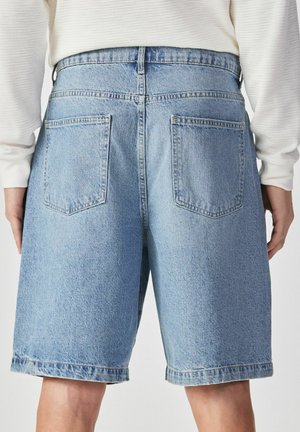 Világoskék farmer rövidnadrág hátul zsebekkel, amelyet egy fehér hosszú ujjú felsőt viselő személy visel, deréktól lefelé látható.