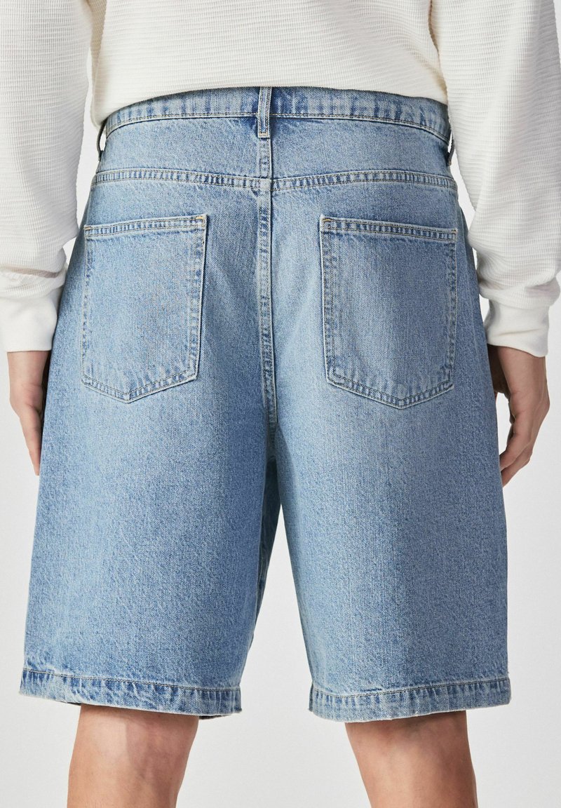 Short en jean bleu clair avec poches arrière porté par une personne vêtue d'un haut blanc à manches longues, vu de la taille vers le bas.