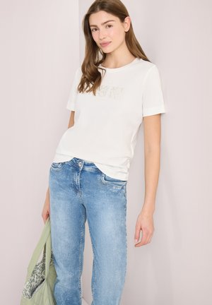 Jeune femme aux longs cheveux bruns portant un t-shirt blanc à manches courtes et un jean bleu clair, tenant un sac vert à motifs.