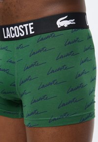 Πράσινο σορτς μποξέρ με μπλε σκουρόχρωμο μοτίβο "Lacoste" και μαύρη μέση με λευκή επιγραφή "LACOSTE" και λογότυπο κροκόδειλου στο ισχίο ενός ατόμου.
