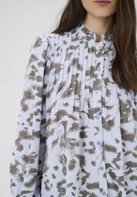 Blusa azzurro chiaro con un motivo astratto marrone, dotata di plissettature, colletto a fascia e chiusura con bottoni. Realizzata in un tessuto morbido e strutturato.