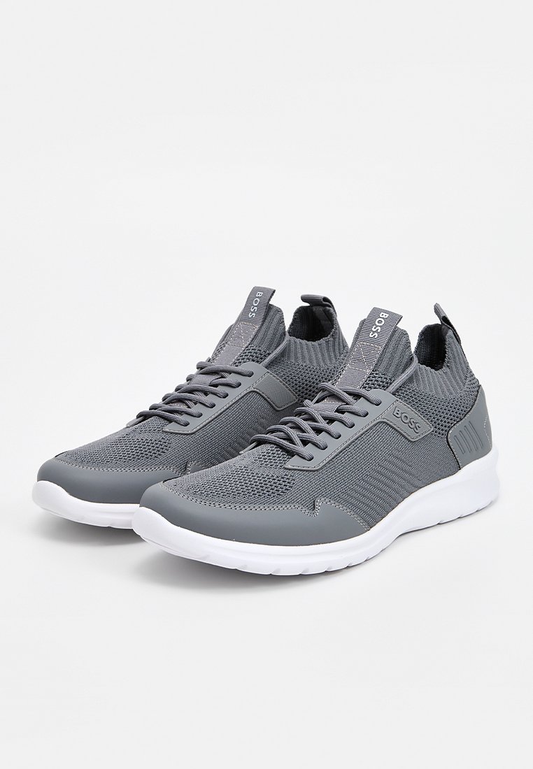 Zapatos deportivos grises con parte superior de tejido transpirable, detalles lisos de goma y suela blanca. Cordones y lengüetas para un ajuste seguro.