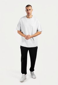 Chemise ample à manches courtes en gris clair, associée à un pantalon noir, présentant un design simple avec une poche frontale. Des baskets blanches complètent le look.
