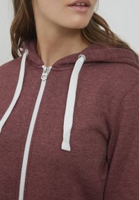 Haut à zip bordeaux fabriqué en tissu doux, avec un cordon blanc, une fermeture éclair sur le devant et une capuche avec un motif simple et texturé.