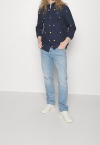 Camisa de manga larga azul marino con patrones coloridos, combinada con pantalones vaqueros de un color azul claro y zapatillas blancas. La camisa tiene un diseño de botones.