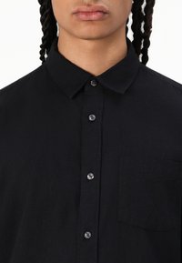 Svart skjorte med knapper laget av teksturert stoff, med klassisk krage, syv knapper og en enkelt brystlomme.