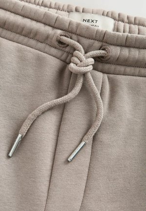Beige joggingbroek tailleband met een gestrikte trekkoordknoop en een zichtbaar merklabel aan de binnenkant van de tailleband met de tekst "NEXT".
