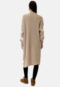 Długi, luźny różowy kardigan z dzianiny z białymi wzorami serc na rękawach, wykończony prążkowanym dołem, noszony na czarnych legginsach i botkach.