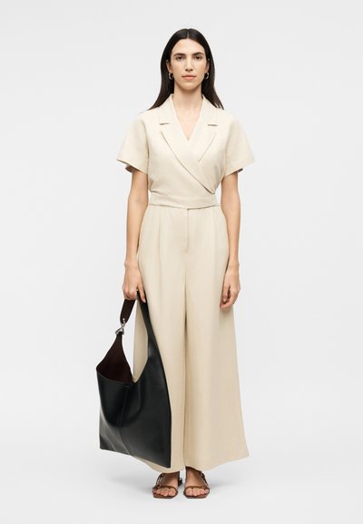 Γυναίκα με μακριά σκούρα μαλλιά, ντυμένη με μπεζ ολόσωμη φόρμα και σανδάλια, κρατώντας μια μεγάλη μαύρη δερμάτινη τσάντα με φαρδιά ταινία.