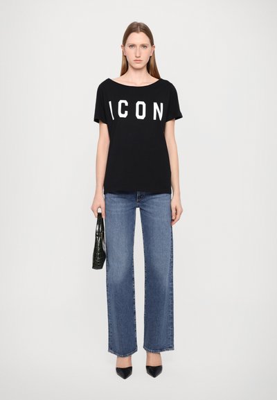 T-shirt noir à manches courtes avec "ICON" imprimé en blanc, associé à un jean large bleu clair et des escarpins noirs à bout pointu. Tenant un sac vert.