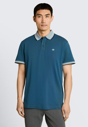 Mężczyzna stoi, ubrany w niebieską koszulkę polo z szarym i białym paskowanym kołnierzykiem i mankietami rękawów, w połączeniu z szarymi spodniami.