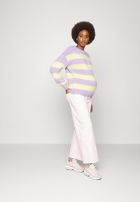Suéter riscado em roxo e amarelo, calças largas rosas e sneakers rosa claro com meias de bolinhas. Modelo exibe uma barriga a crescer.