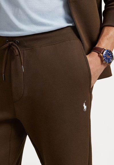 Pantalones de chándal marrones con cintura ajustable, que presentan un logo bordado blanco en el muslo y una tela suave y texturizada.