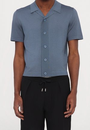 Mann trägt ein blaues Hemd mit kurzen Ärmeln und Knöpfen, mit Kragen, kombiniert mit schwarzen Hosen mit Kordelzug.