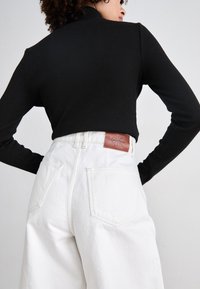 Zwarte geribbelde top met lange mouwen, gecombineerd met hoge witte denimbroeken. Leren merklabel zichtbaar op de achterkant van de tailleband.