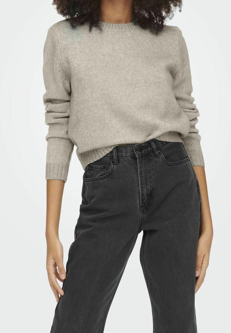 Pull beige en maille avec un col rond et des manches longues, associé à un jean noir taille haute. Le tissu présente un motif texturé.