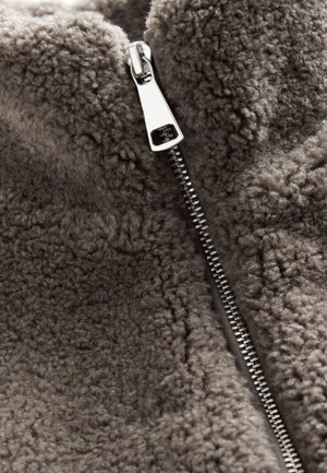 Veste en polaire en tissu gris doux avec une surface texturée. Dotée d'une fermeture éclair argentée sur le devant.