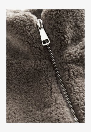 Veste en polaire en tissu gris doux avec une surface texturée. Dotée d'une fermeture éclair argentée sur le devant.