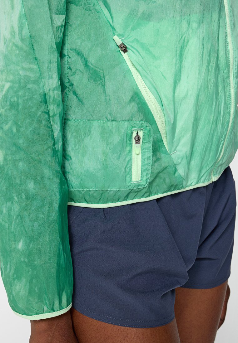 Lekka, półprzezroczysta zielona kurtka z zapięciem na zamek i kieszenią. Kontrastowy zamek i wykończenia w mięcie. Połączona z granatowymi szortami.