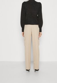 Osoba ubrana w beżowe, szerokie spodnie cargo i czarny sweter z długim rękawem, stojąca plecami do kamery na jednolitym tle.