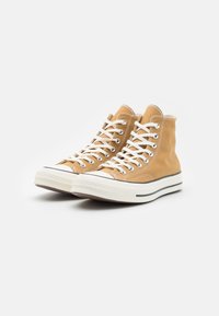 Zapatillas altas hechas de lona marrón con cordones blancos, suelas de goma y acentos en negro. Superficie texturizada con detalles de costura en contraste.