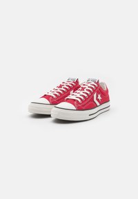 Une paire de Converse All Star rouge à tige basse avec des lacets blancs, des embouts en caoutchouc et le logo étoile sur le côté, sur un fond blanc.