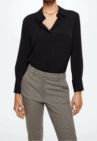 Blouse noire à manches longues avec un col, associée à un pantalon taille haute à motif pied de poule en beige et noir.