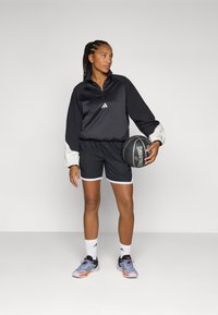 Svart treningsjakke med hvite detaljer, kombinert med svarte shorts og grå treningsko. Holder en basketball, står på en ensfarget bakgrunn.